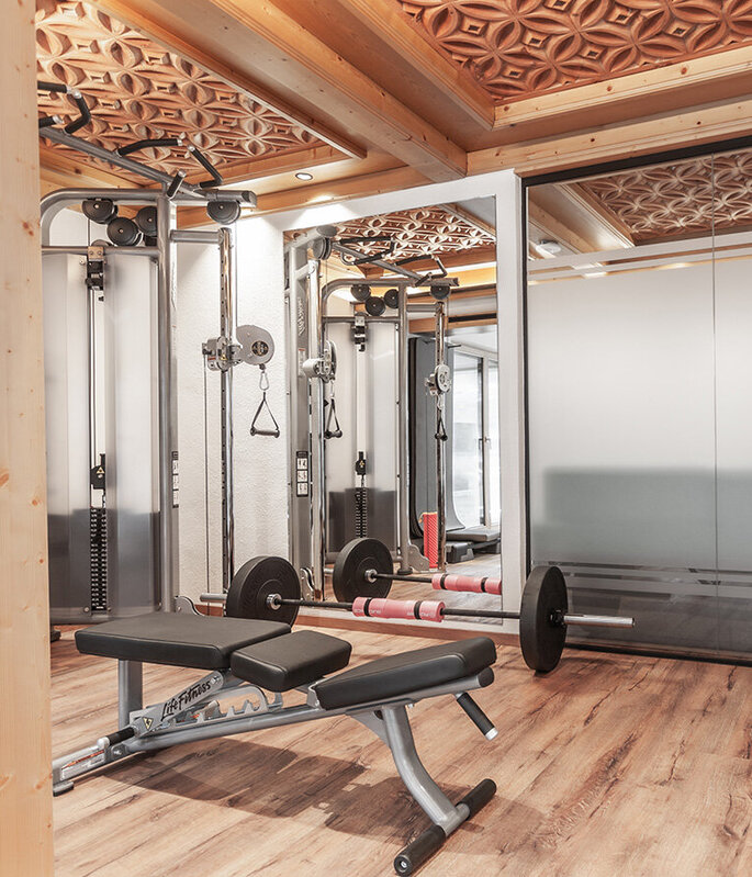 Ein moderner Fitnessraum mit Hantelbank, Langhantel und Fitnessgeräten, ausgestattet mit Holzböden und einer kunstvoll verzierten Holzdecke sowie Wänden aus Milchglas.