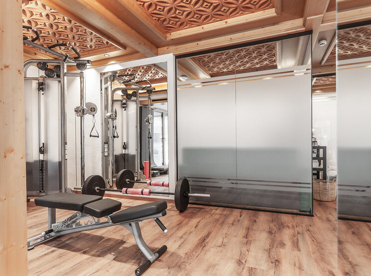 Ein moderner Fitnessraum mit Hantelbank, Langhantel und Fitnessgeräten, ausgestattet mit Holzböden und einer kunstvoll verzierten Holzdecke sowie Wänden aus Milchglas.