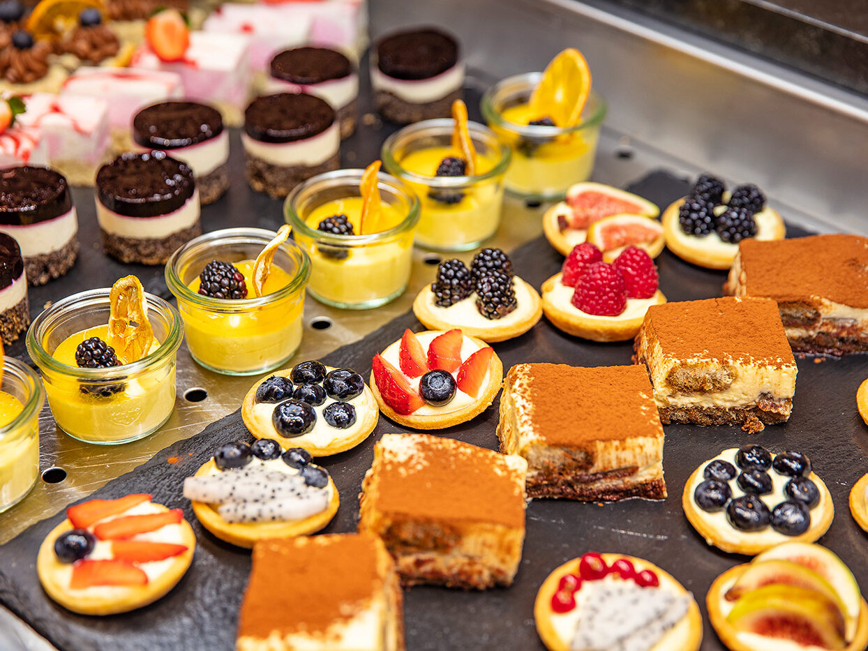 Eine Auswahl an Desserts, darunter Tiramisu-Quadrate, mit Beeren garnierte Obsttörtchen, mehrschichtige Parfaits in Gläsern und Puddingbecher mit Brombeeren und Orangenscheiben, die auf einem Buffettisch angerichtet sind.