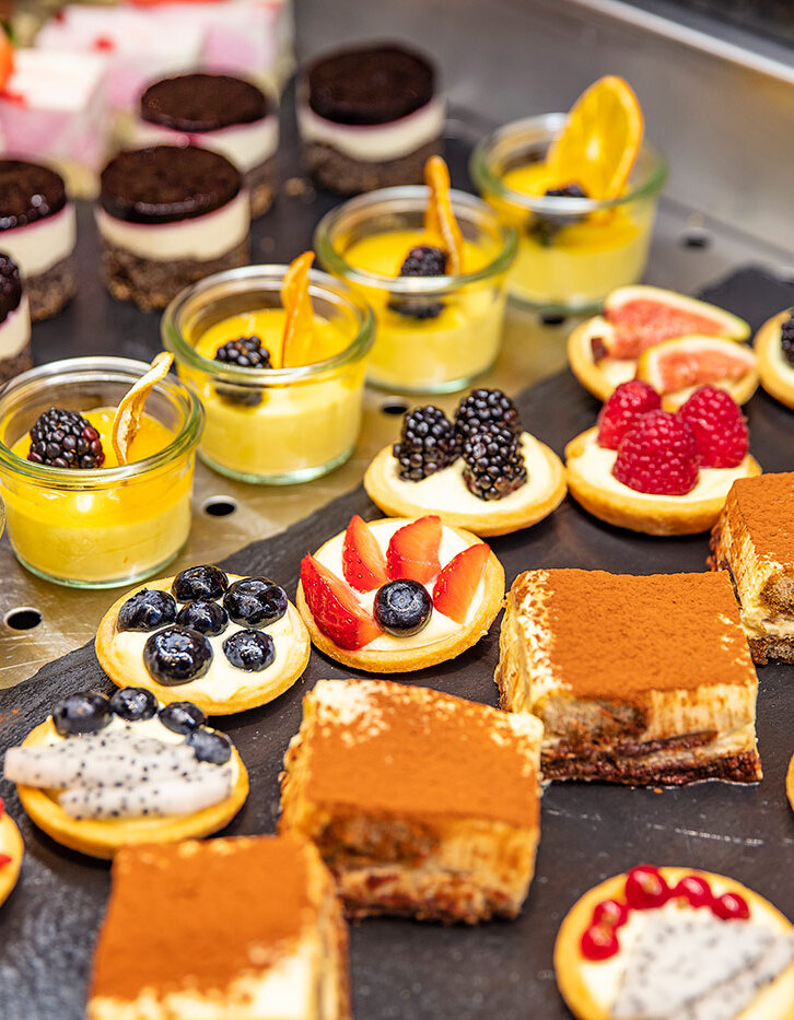 Eine Auswahl an Desserts, darunter Tiramisu-Quadrate, mit Beeren garnierte Obsttörtchen, mehrschichtige Parfaits in Gläsern und Puddingbecher mit Brombeeren und Orangenscheiben, die auf einem Buffettisch angerichtet sind.