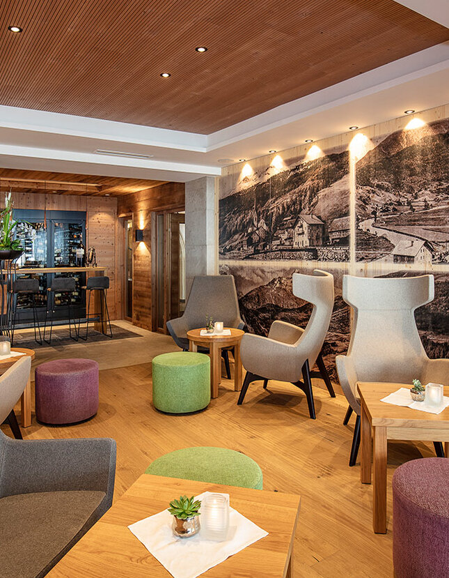 Moderne Lounge mit grauen Sesseln, kleinen Holztischen und bunten Sitzkissen. Eine große Schwarz-Weiß-Wandmalerei ziert eine Wand. Gemütliche Beleuchtung und im Hintergrund ein Teilblick auf einen Barbereich.