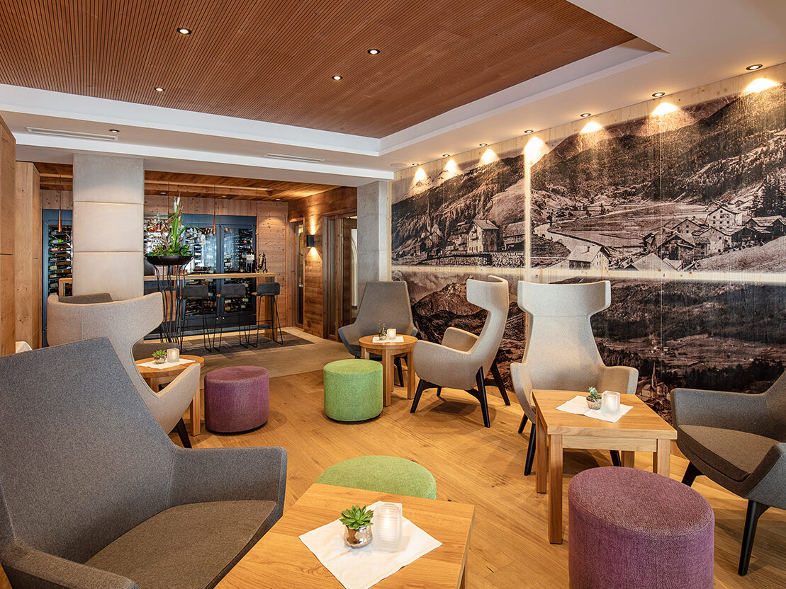 Moderne Lounge mit grauen Sesseln, kleinen Holztischen und bunten Sitzkissen. Eine große Schwarz-Weiß-Wandmalerei ziert eine Wand. Gemütliche Beleuchtung und im Hintergrund ein Teilblick auf einen Barbereich.