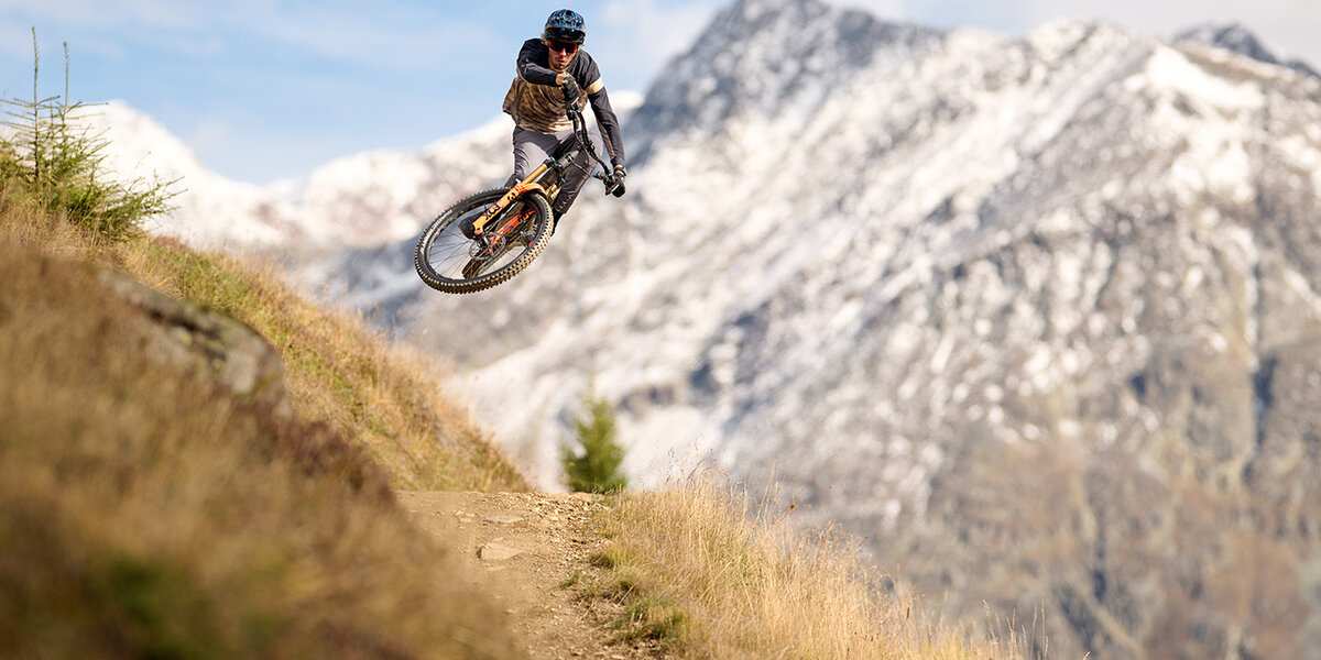 Ein Mountainbiker springt mitten in der Luft von einem Feldweg auf einen grasbewachsenen Hang, im Hintergrund sind schneebedeckte Berge und ein teilweise bewölkter Himmel zu sehen.