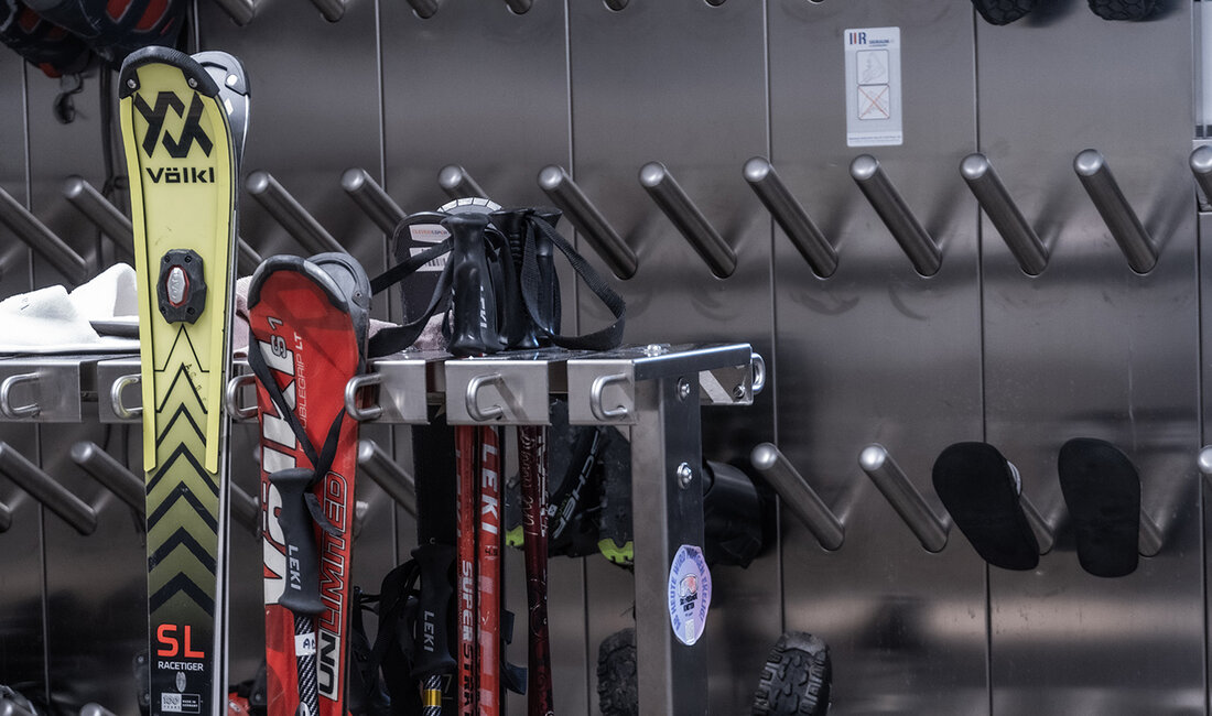 Die Skiausrüstung, darunter Stiefel, Stöcke und Skier, wird in einem Metallregal und an Wandhaltern aufbewahrt, wobei ein Teil der Ausrüstung zum Trocknen aufgehängt ist. Die Szene wirkt ordentlich und zweckmäßig.