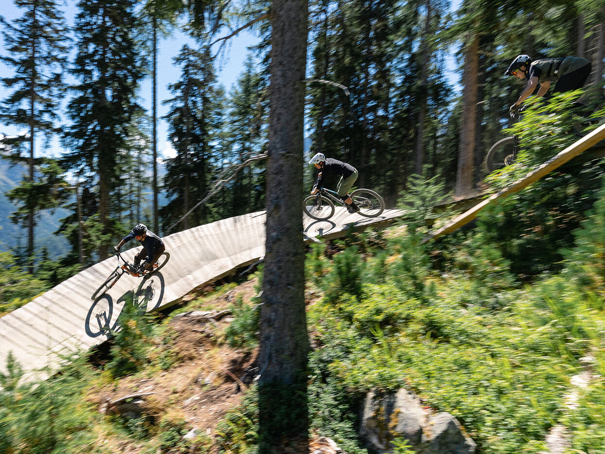 An einem sonnigen Tag fahren drei Mountainbiker einen kurvenreichen Holzpfad hinunter, der durch einen bewaldeten Hang führt, umgeben von hohen Bäumen und viel Grün.
