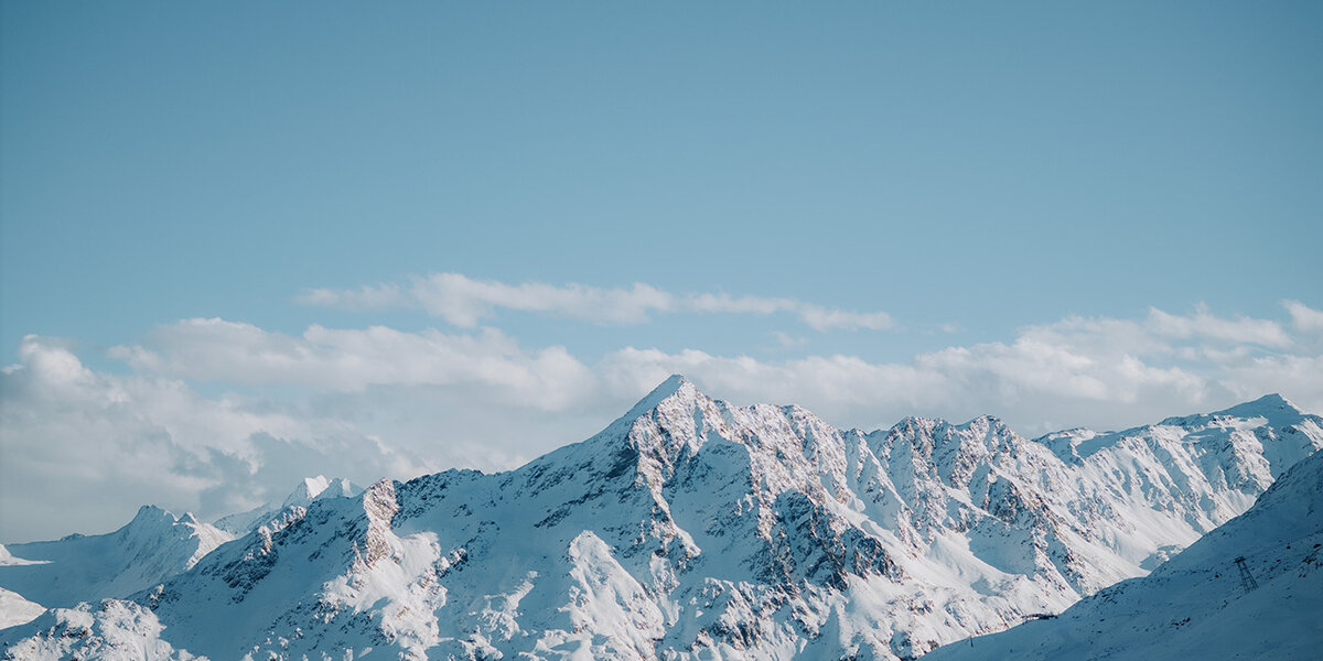 Schneebedeckte Berggipfel unter einem strahlend blauen Himmel, mit sanften Wolken im Hintergrund und Sonnenlicht, das die Bergrücken und Hänge zum Leuchten bringt.