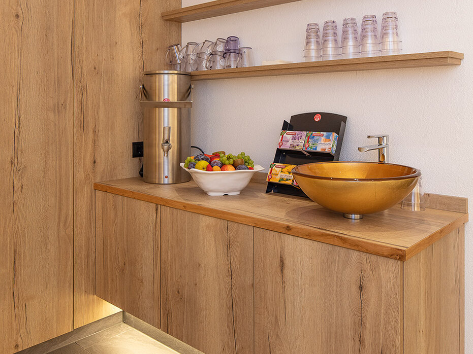 Moderne Teebar mit Holzschränken, einer goldenen Spüle, einer Obstschale, einer Snackauslage, Gläsern, Flaschen und einer Unterbauleuchte. Auf zwei Holzregalen stehen Tassen und Getränke. Das Design ist minimalistisch und hell.
