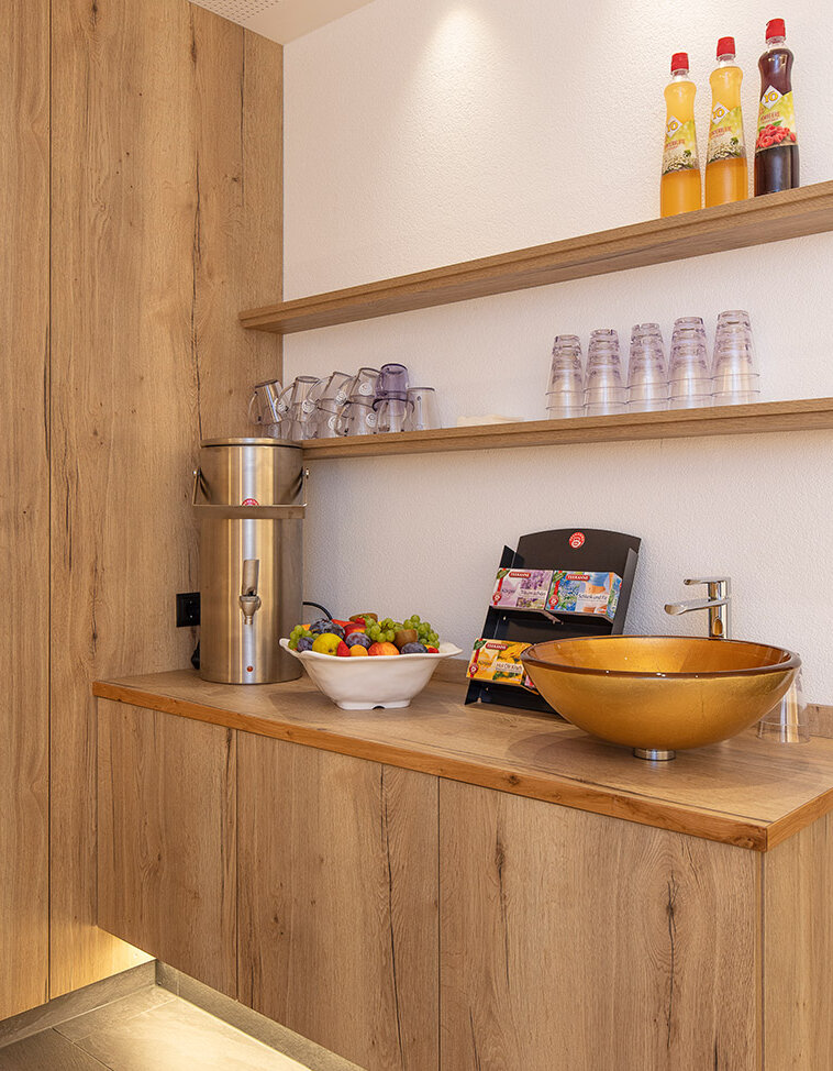 Moderne Teebar mit Holzschränken, einer goldenen Spüle, einer Obstschale, einer Snackauslage, Gläsern, Flaschen und einer Unterbauleuchte. Auf zwei Holzregalen stehen Tassen und Getränke. Das Design ist minimalistisch und hell.