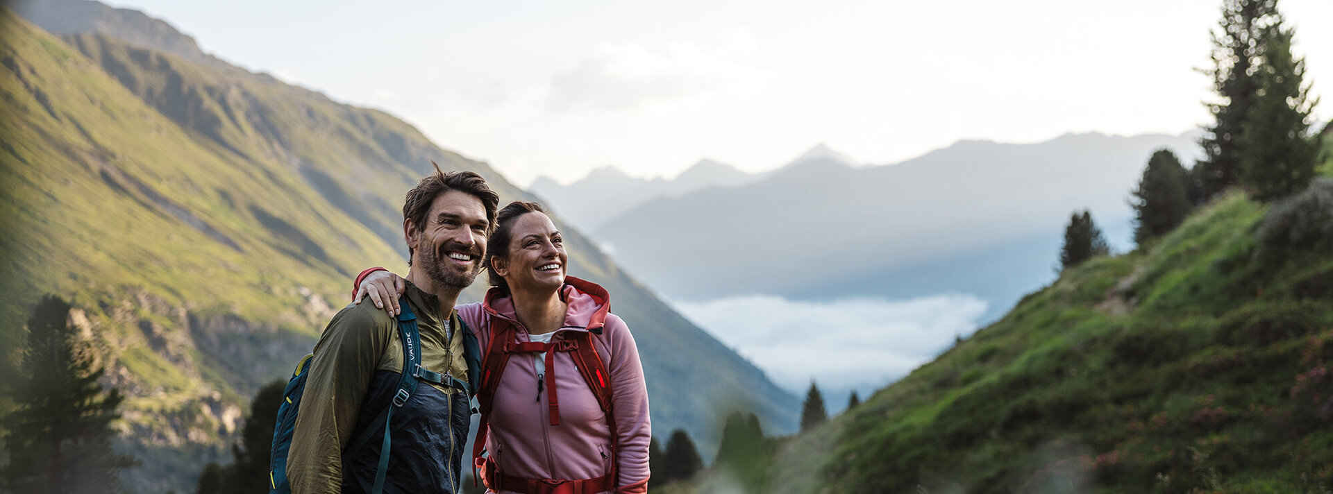 Ein Mann und eine Frau mit Rucksäcken stehen lächelnd nebeneinander auf einem grünen Bergpfad, umgeben von Hügeln und Bäumen, im Hintergrund sind nebelverhangene Berge zu sehen.