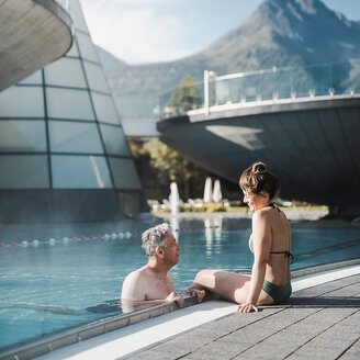 Ein Mann und eine Frau entspannen sich am Rand eines Außenpools im Aqua Dome, im Hintergrund sind moderne, futuristische Gebäude und ein malerischer Berg zu sehen.