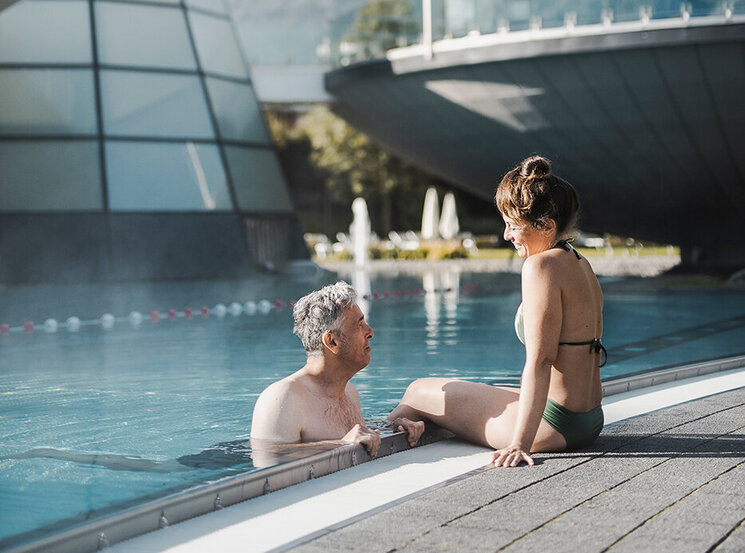 Ein Mann und eine Frau entspannen sich am Rand eines Außenpools im Aqua Dome, im Hintergrund sind moderne, futuristische Gebäude und ein malerischer Berg zu sehen.