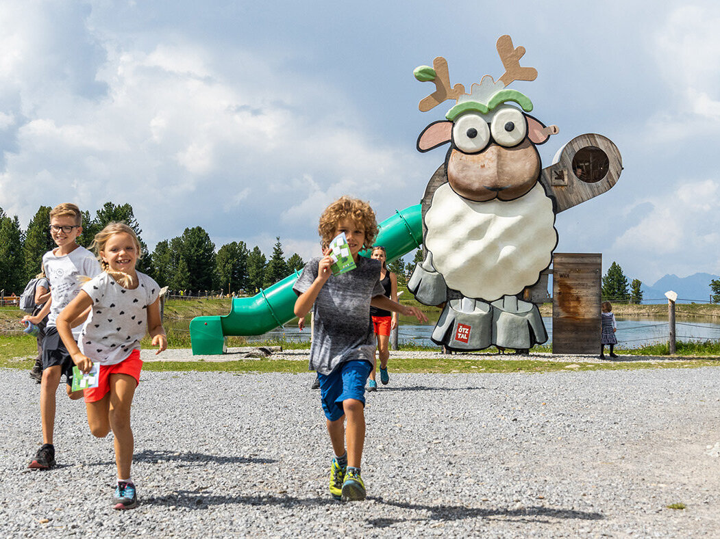 Drei Kinder rennen auf einem Schotterweg in der Nähe einer verspielten Spielplatzrutsche, die wie ein Schaf mit Brille geformt ist. Im Hintergrund sind Bäume, Gras und ein See unter einem teilweise bewölkten Himmel zu sehen.