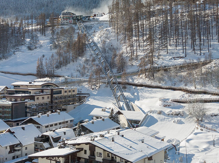 Ein verschneites Bergdorf mit Häusern, geparkten Autos und schneebedeckten Bäumen. Der Zentrum Shuttle führt hinter dem Hotel Hubertus den Hang hinauf, im Hintergrund sind Berge und ein strahlend blauer Himmel zu sehen.
