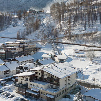 Ein verschneites Bergdorf mit Häusern, geparkten Autos und schneebedeckten Bäumen. Der Zentrum Shuttle führt hinter dem Hotel Hubertus den Hang hinauf, im Hintergrund sind Berge und ein strahlend blauer Himmel zu sehen.