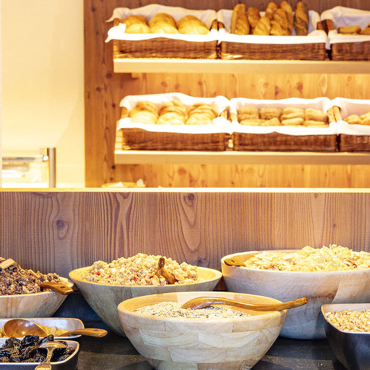 Im Vordergrund ein Frühstücksbuffet mit Holzschalen voller Müsli, Trockenfrüchten und Nüssen, im Hintergrund Regale mit verschiedenen Brotsorten und Gebäck in Körben.