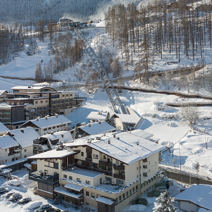 Ein schneebedecktes Dorf mit mehreren Gebäuden und geparkten Autos, das am Fuße eines von Bäumen gesäumten, bewaldeten Hügels liegt. Im Hintergrund erheben sich schneebedeckte Berge unter einem strahlend blauen Himmel. Zu sehen ist die Außenansicht des Hotels und der Zentrum Shuttle gleich dahinter.