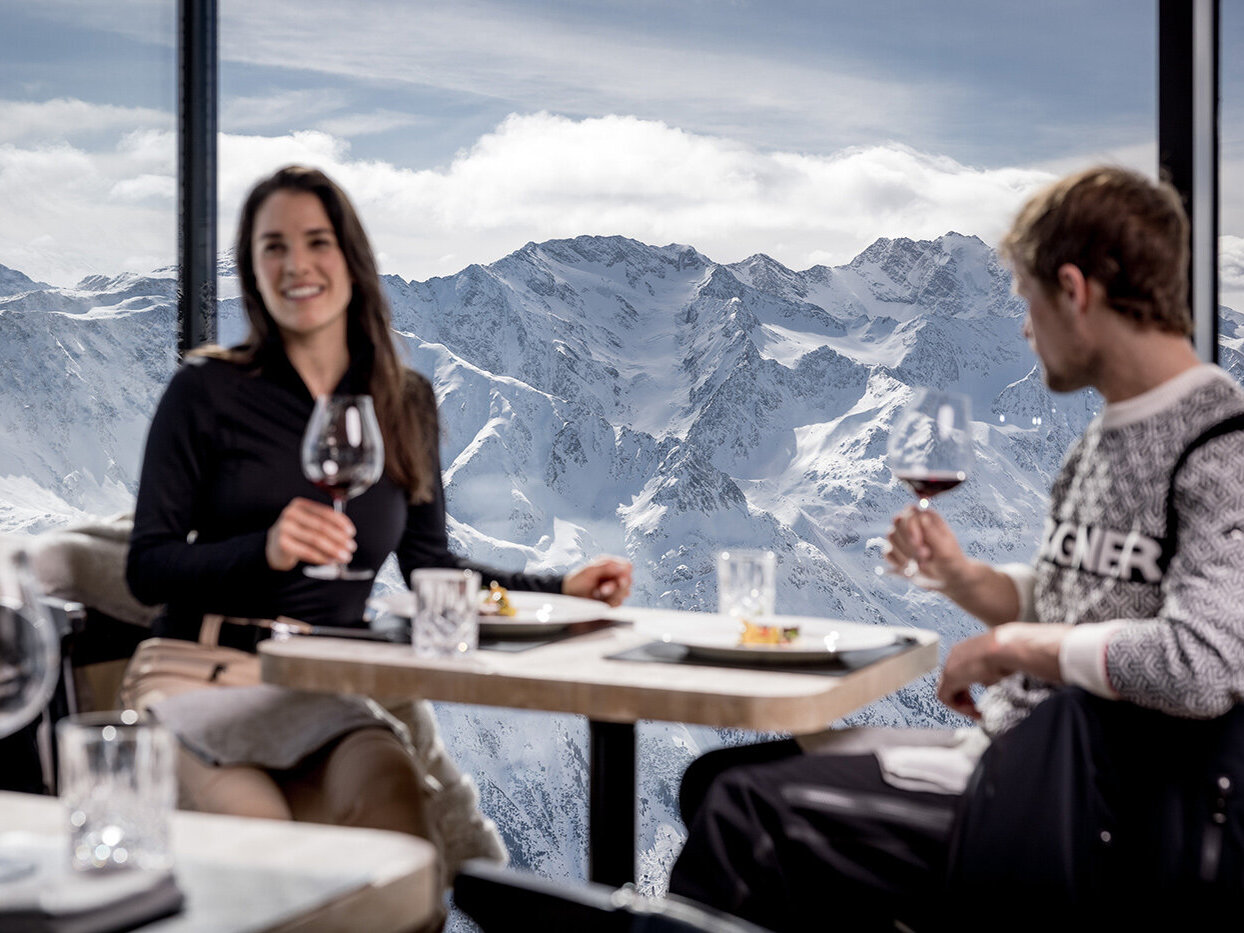 Zwei Personen sitzen an einem Tisch in einem Restaurant am Fenster und halten Weingläser in der Hand; im Hintergrund sind unter einem bewölkten Himmel schneebedeckte Berggipfel zu sehen.