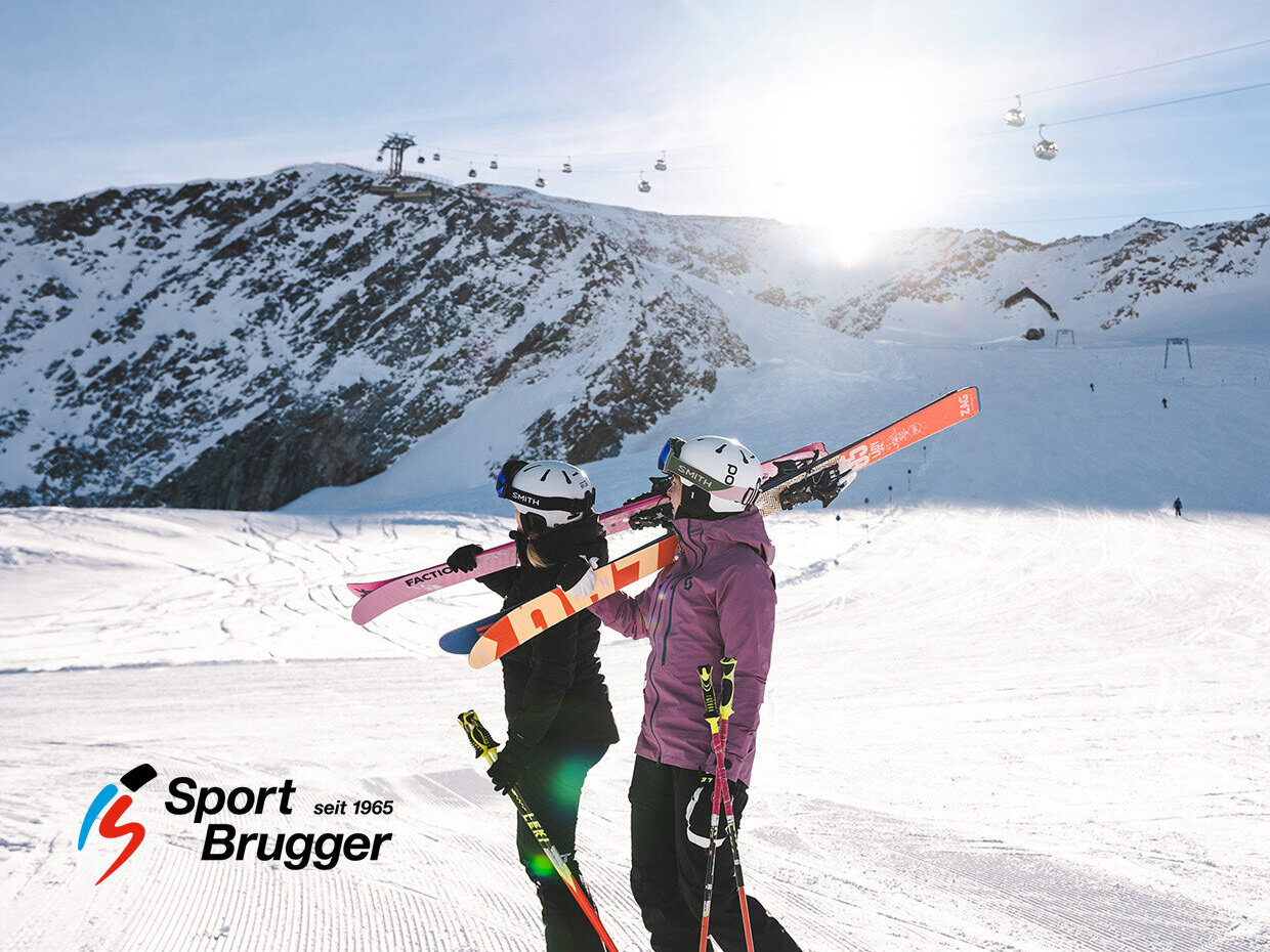Zwei Skifahrer in Winterkleidung steigen mit ihren Skiern auf den Schultern einen verschneiten Hang hinauf, im Hintergrund sind Skilifte und strahlender Sonnenschein zu sehen. In der unteren linken Ecke ist das Logo von Sport Brugger zu sehen.