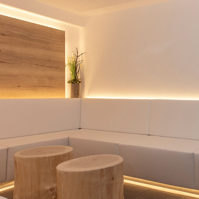 Eine moderne, minimalistische Infrarot-Lounge mit weißen Polsterkissen, zwei runden Holzhockern, einer kleinen Pflanze in der Ecke und sanfter Stimmungsbeleuchtung entlang der Wand.