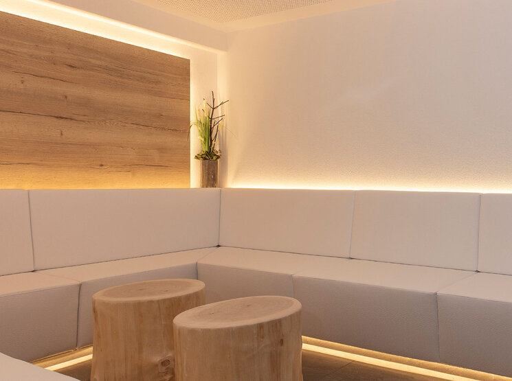 Eine moderne, minimalistische Infrarot-Lounge mit weißen Polsterkissen, zwei runden Holzhockern, einer kleinen Pflanze in der Ecke und sanfter Stimmungsbeleuchtung entlang der Wand.