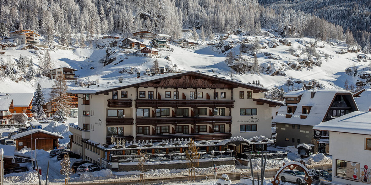 Ein verschneites Bergdorf mit Gebäuden im Chalet-Stil, schneebedeckten Kiefern und einem hohen Berggipfel im Hintergrund unter strahlend blauem Himmel. Zu sehen ist die Außenansicht vom Hotel Hubertus.