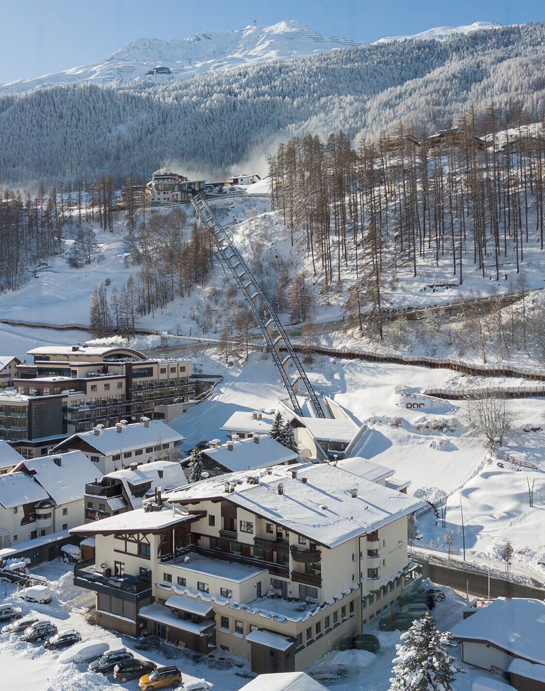 Ein verschneites Bergdorf mit Häusern, geparkten Autos und schneebedeckten Bäumen. Der Zentrum Shuttle führt hinter dem Hotel Hubertus den Hang hinauf, im Hintergrund sind Berge und ein strahlend blauer Himmel zu sehen.