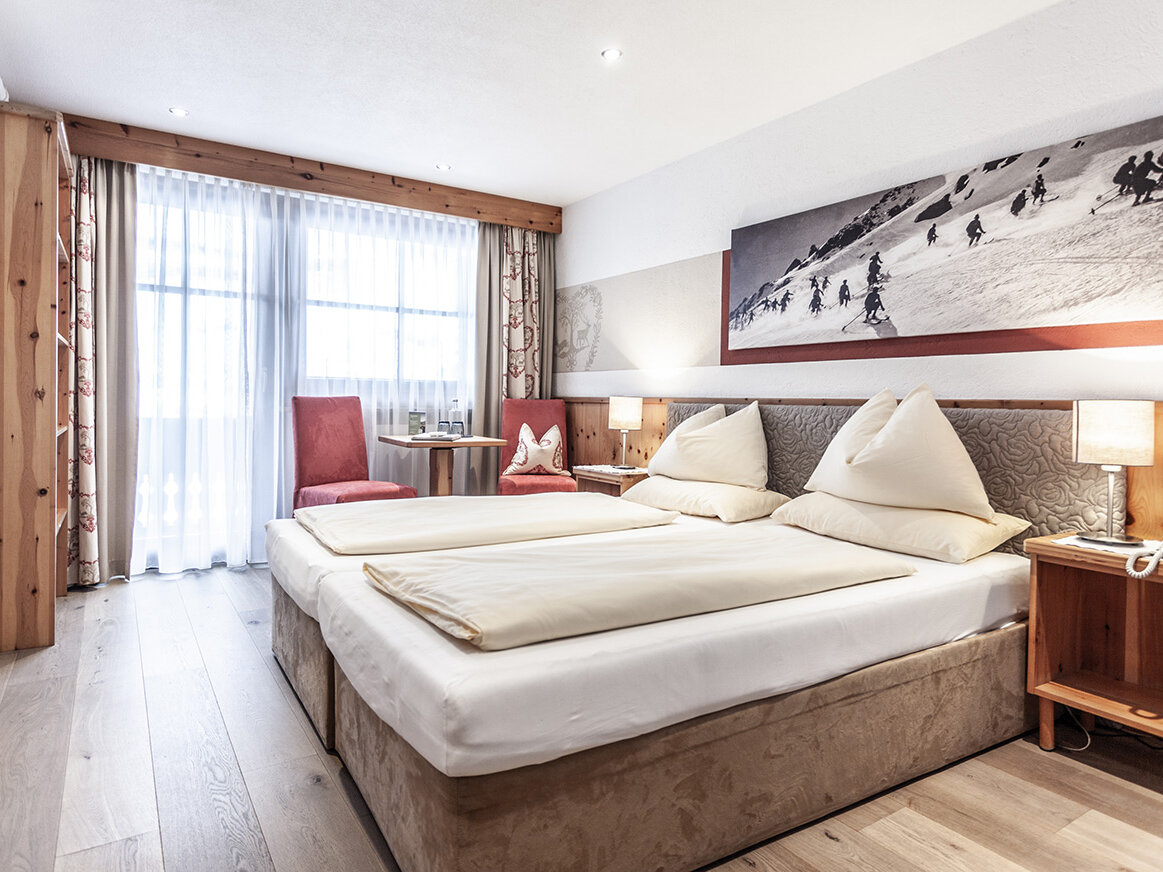 Ein helles Hotelzimmer mit einem Doppelbett, Holzmöbeln, einem kleinen Tisch mit zwei roten Stühlen, einem großen Fenster mit transparenten Vorhängen und einem schwarz-weißen Skifoto über dem Bett.