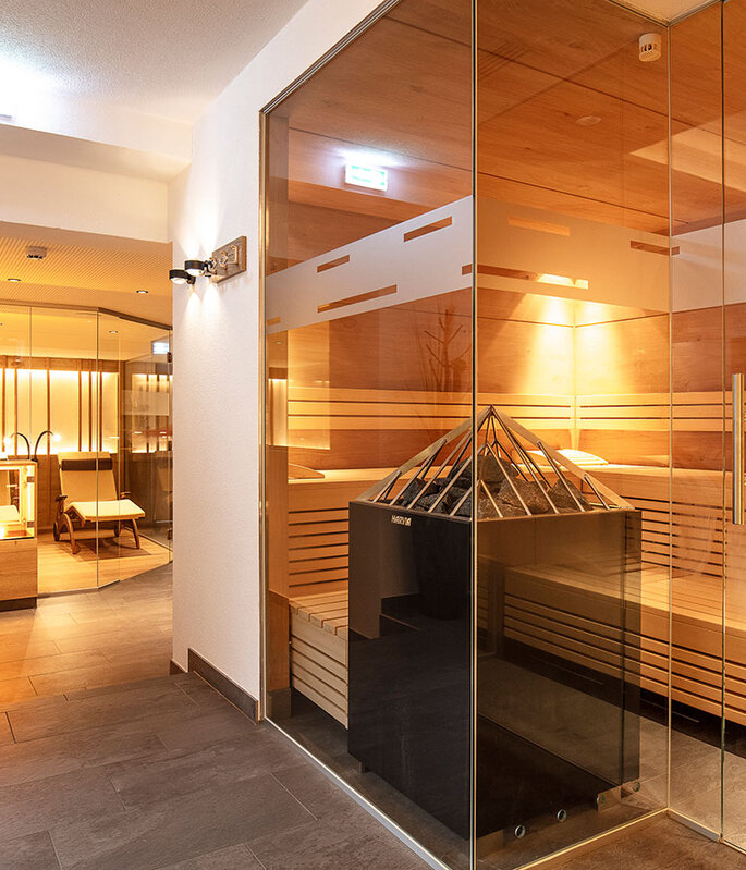 Ein moderner Wellnessbereich mit einer großen, verglasten Biosauna, Holzbänken im Inneren, warmer Beleuchtung und einer Wellness-Dekoration, zu der unter anderem eine hohe Vase mit Pflanzen gehört, die eine ruhige und entspannende Atmosphäre schafft.