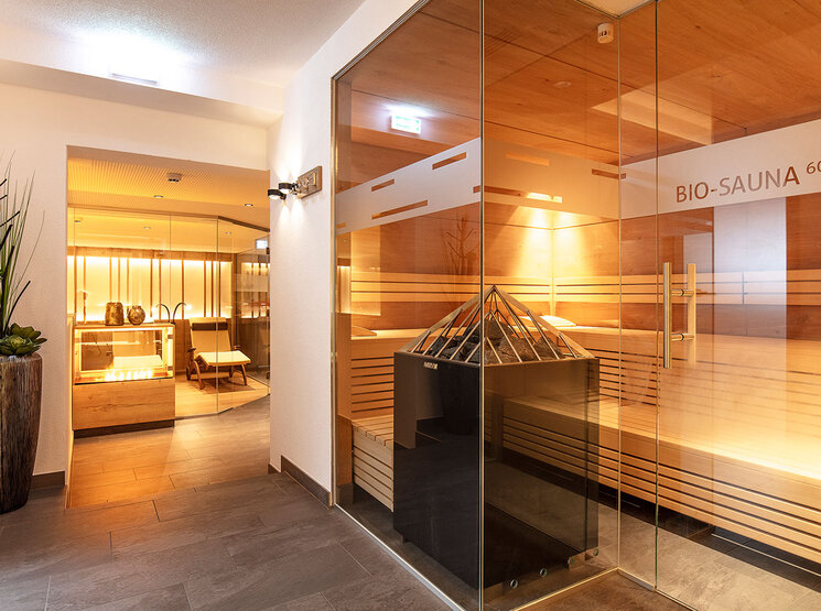 Ein moderner Wellnessbereich mit einer großen, verglasten Biosauna, Holzbänken im Inneren, warmer Beleuchtung und einer Wellness-Dekoration, zu der unter anderem eine hohe Vase mit Pflanzen gehört, die eine ruhige und entspannende Atmosphäre schafft.