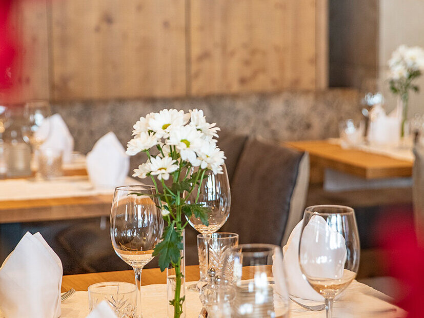 Eine Nahaufnahme eines Restauranttisches, gedeckt mit weißen Servietten, Weingläsern, Besteck und einer Vase mit weißen Blumen. Im Hintergrund sind weitere Tische und Holzdekorationen zu sehen, im Vordergrund sind rote Blumen verschwommen abgebildet.