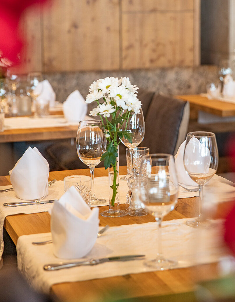 Eine Nahaufnahme eines Restauranttisches, gedeckt mit weißen Servietten, Weingläsern, Besteck und einer Vase mit weißen Blumen. Im Hintergrund sind weitere Tische und Holzdekorationen zu sehen, im Vordergrund sind rote Blumen verschwommen abgebildet.