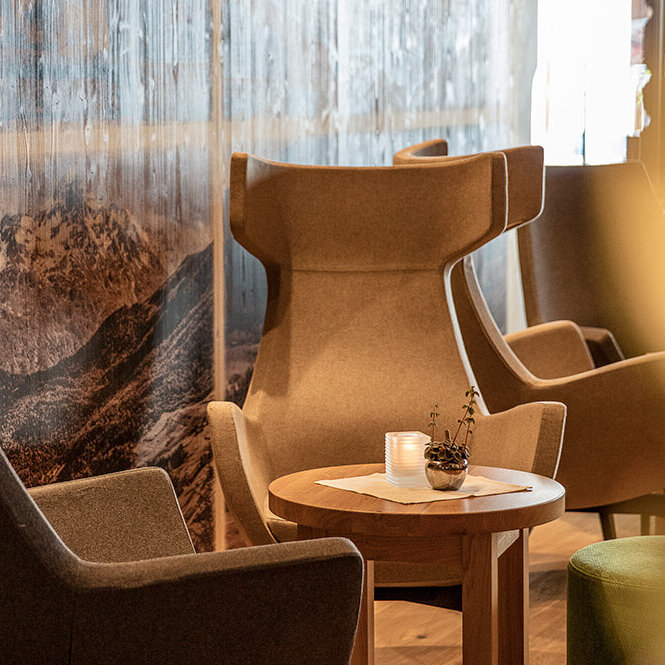 Zwei moderne Sessel und ein runder Holztisch mit einer Kerze stehen neben einer Wand, die mit einem Bergmotiv verziert ist, und schaffen so eine gemütliche, einladende Lounge-Atmosphäre.