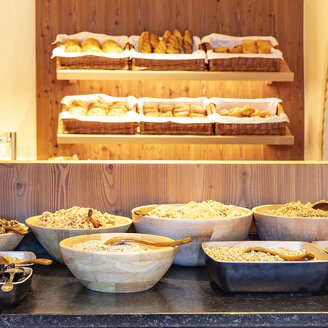 Im Vordergrund ein Frühstücksbuffet mit Holzschalen voller Müsli, Trockenfrüchten und Nüssen, im Hintergrund Regale mit verschiedenen Brotsorten und Gebäck in Körben.