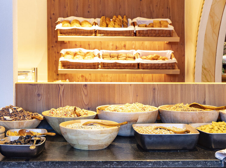 Im Vordergrund ein Frühstücksbuffet mit Holzschalen voller Müsli, Trockenfrüchten und Nüssen, im Hintergrund Regale mit verschiedenen Brotsorten und Gebäck in Körben.