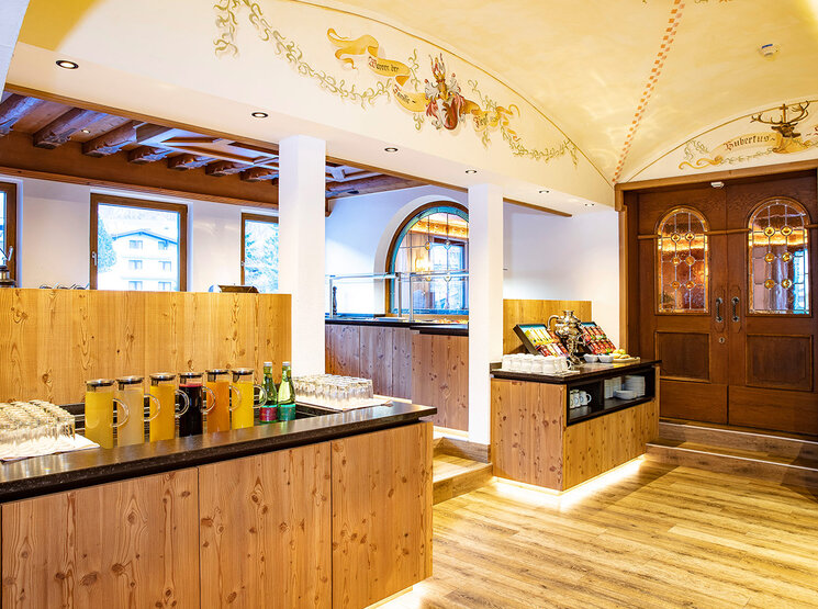 In einer gemütlichen Cafeteria mit Holztheken werden Getränke und Snacks angeboten. Warme Beleuchtung, dekorative Deckenmalereien und Bogenfenster schaffen eine einladende Atmosphäre. Im Hintergrund ist eine Holztür mit Buntglas zu sehen.