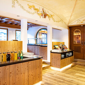 In einer gemütlichen Cafeteria mit Holztheken werden Getränke und Snacks angeboten. Warme Beleuchtung, dekorative Deckenmalereien und Bogenfenster schaffen eine einladende Atmosphäre. Im Hintergrund ist eine Holztür mit Buntglas zu sehen.