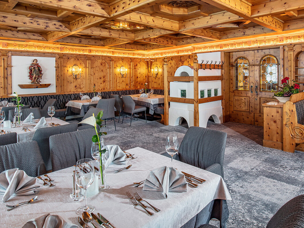 Ein elegantes Restaurant mit Holzwänden und -decke, grauem Teppichboden, ordentlich gedeckten Tischen mit weißen Tischdecken, gefalteten Servietten, Weingläsern, grauen Stühlen, Wandbildern und dekorativer Beleuchtung, die eine gemütliche, gehobene Atmosphäre schaffen.