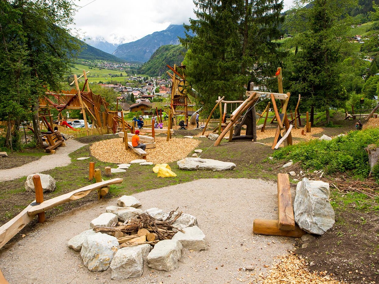 Ein Spielplatz aus Holz mit Schaukeln, Rutschen und Bänken inmitten einer üppig grünen, bergigen Landschaft. Kinder und Erwachsene spielen und entspannen sich. Im Vordergrund sind eine Feuerstelle und Wege zu sehen, im Hintergrund sind Bäume und das Dorf zu erkennen.