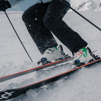 Nahaufnahme der Beine und Skier eines Skifahrers, der durch Neuschnee gleitet, wobei Schnee hochspritzt. Der Skifahrer trägt eine schwarze Hose und weiße Skischuhe. Im Hintergrund ist eine verschneite Berglandschaft zu sehen.