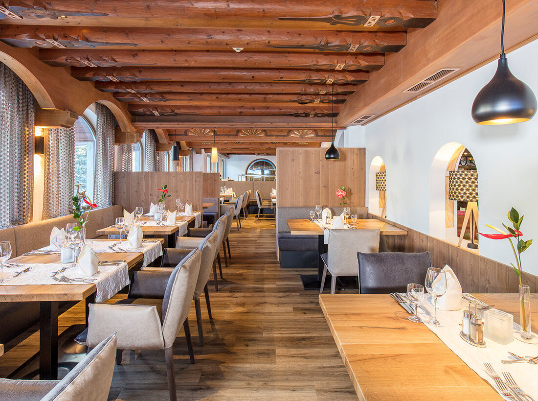Ein gemütlicher Restaurantbereich mit Holzböden, Deckenbalken, großen Fenstern, ordentlich gedeckten Tischen mit weißen Servietten und modernen Hängeleuchten. Auf einigen Tischen stehen einzelne rote Blumen in Vasen.
