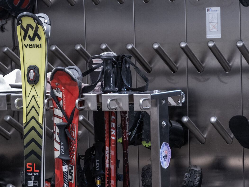 Die Skiausrüstung, darunter Skier, Skistöcke, Skischuhe und ein Helm, wird in einem Abstell- oder Trockenraum an einem Metallregal und an Wandhaken aufbewahrt.