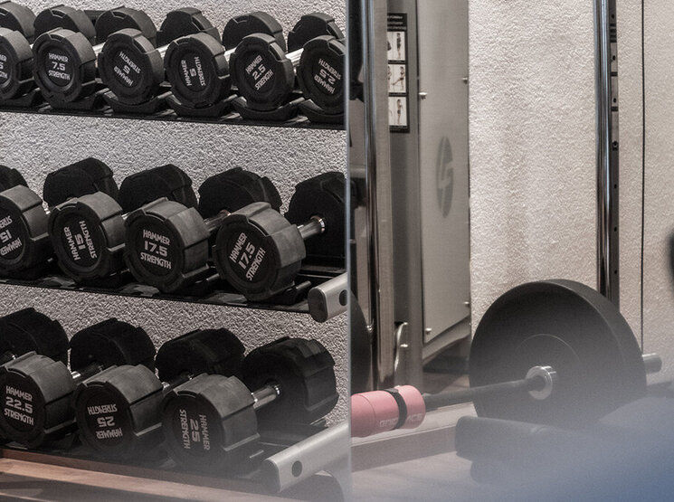 Ein Fitnessbereich mit einem Hantelständer, einem Krafttrainingsgerät und einer Langhantel mit Hantelscheiben auf dem Boden. Die Geräte sind ordentlich angeordnet, und die Umgebung wirkt sauber und modern.
