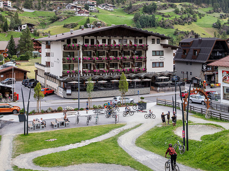 Außenansicht vom Hotel Hubertus mit roten Blumen auf den Balkonen in einem Bergdorf, umgeben von grünen Hügeln und Kiefernwäldern. Im Vordergrund fahren Radfahrer im Bikepark durch kurvenreiche Wege. Im Hintergrund sind Berggipfel und ein bewölkter Himmel zu sehen.