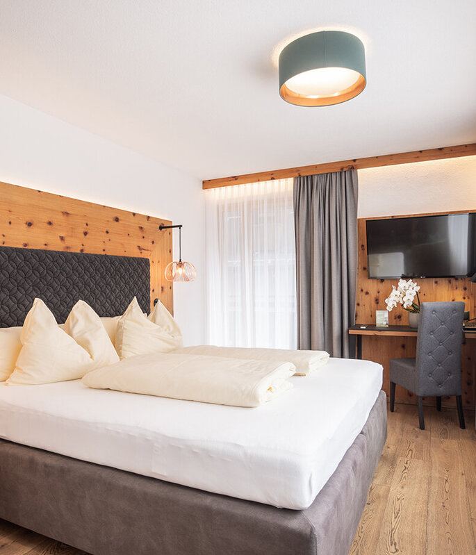 Modernes Hotelzimmer mit einem großen Bett, cremefarbener Bettwäsche, holzvertäfelten Wänden, Hängeleuchten, einem Schreibtisch mit Stuhl, einem Fernseher und großen Fenstern mit transparenten Vorhängen, durch die Tageslicht hereinströmt.