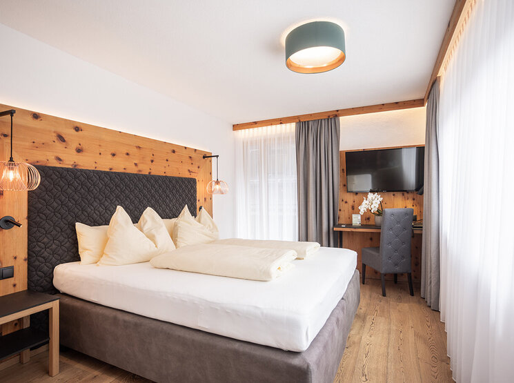 Modernes Hotelzimmer mit einem großen Bett, cremefarbener Bettwäsche, holzvertäfelten Wänden, Hängeleuchten, einem Schreibtisch mit Stuhl, einem Fernseher und großen Fenstern mit transparenten Vorhängen, durch die Tageslicht hereinströmt.