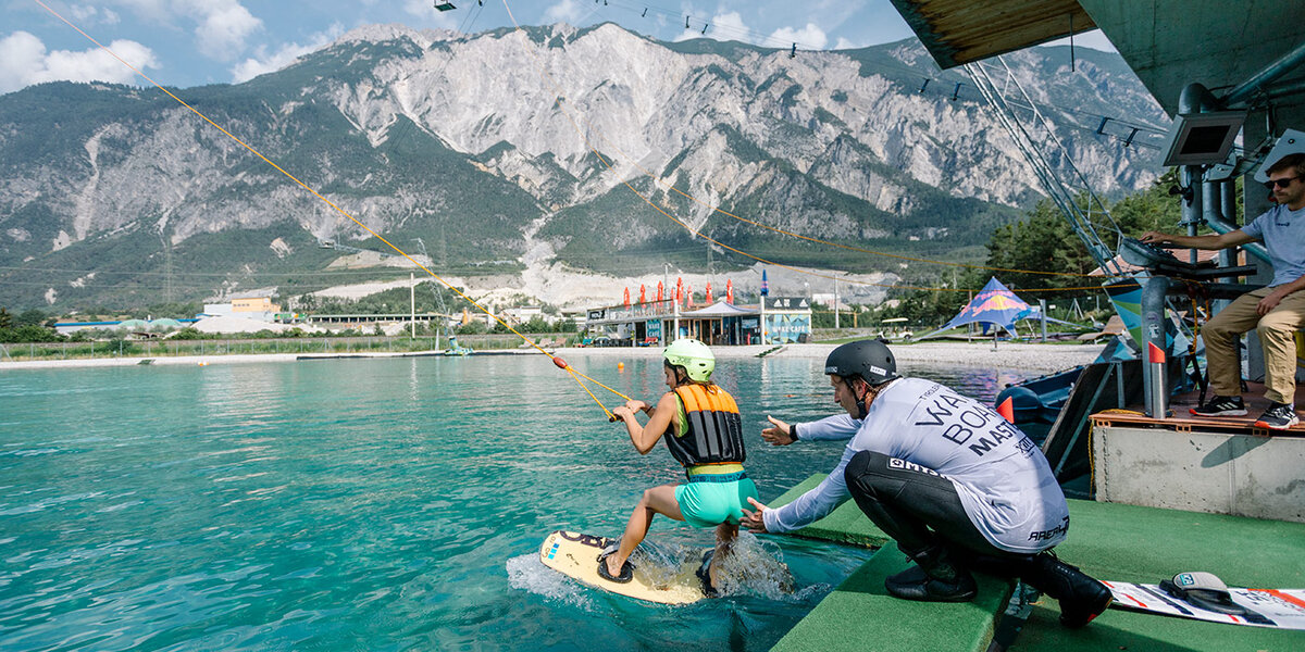 Eine Person auf einem Wakeboard in der Area 47 wird von einer Seilbahn über türkisfarbenes Wasser gezogen, während ein Lehrer in der Nähe kniet. Im Hintergrund sind Berge und Gebäude zu sehen.