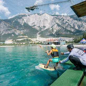 Eine Person auf einem Wakeboard in der Area 47 wird von einer Seilbahn über türkisfarbenes Wasser gezogen, während ein Lehrer in der Nähe kniet. Im Hintergrund sind Berge und Gebäude zu sehen.