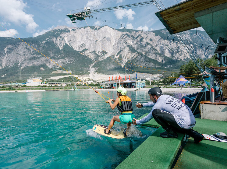 Eine Person auf einem Wakeboard in der Area 47 wird von einer Seilbahn über türkisfarbenes Wasser gezogen, während ein Lehrer in der Nähe kniet. Im Hintergrund sind Berge und Gebäude zu sehen.