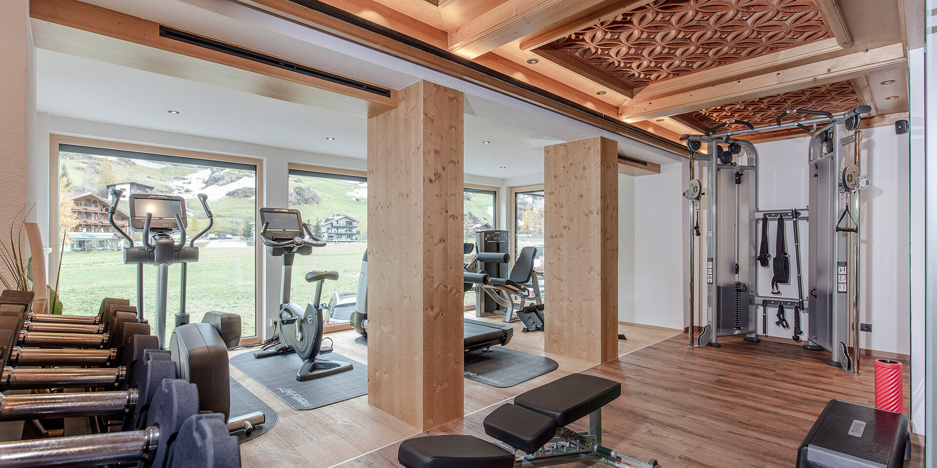Modernes Fitnessstudio mit Holzelementen, ausgestattet mit Hanteln, Trainingsbänken, Cardiogeräten vor großen Fenstern und einer multifunktionalen Kabelzugmaschine. Durch die Glasfronten strömt viel Tageslicht herein und bietet einen malerischen Blick ins Freie.