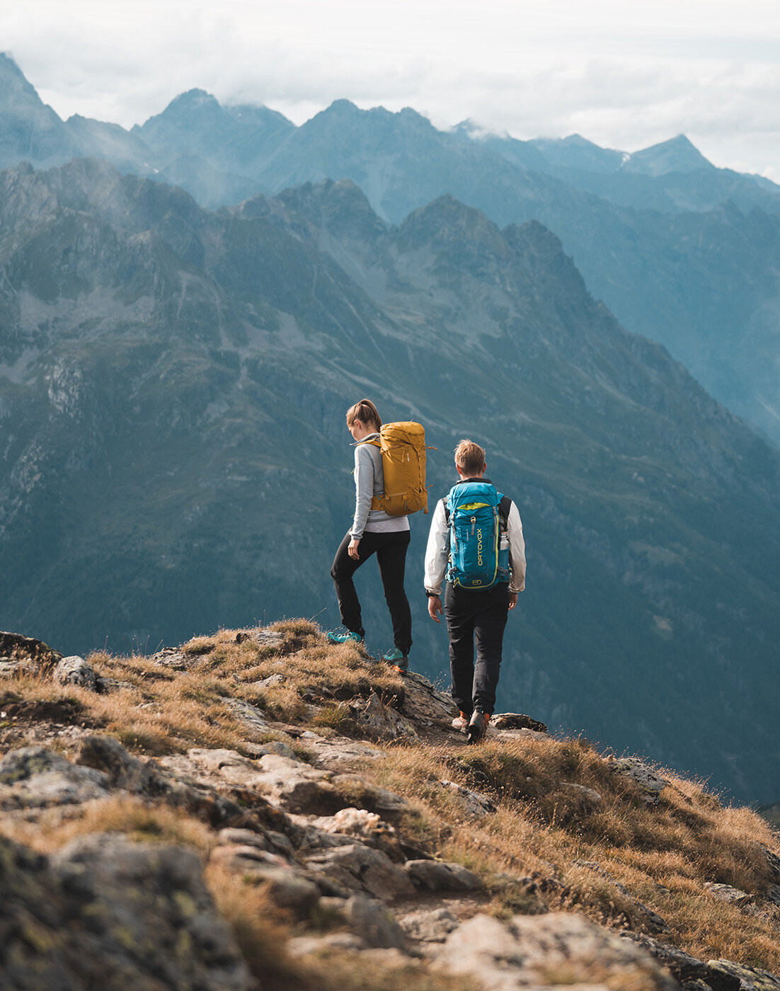 Zwei Wanderer mit Rucksäcken stehen auf einem felsigen Bergpfad und blicken unter bewölktem Himmel auf eine Kette entfernter Gipfel. Die Szene liegt in erhöhter Lage und ist von schroffem, malerischem Gelände umgeben.